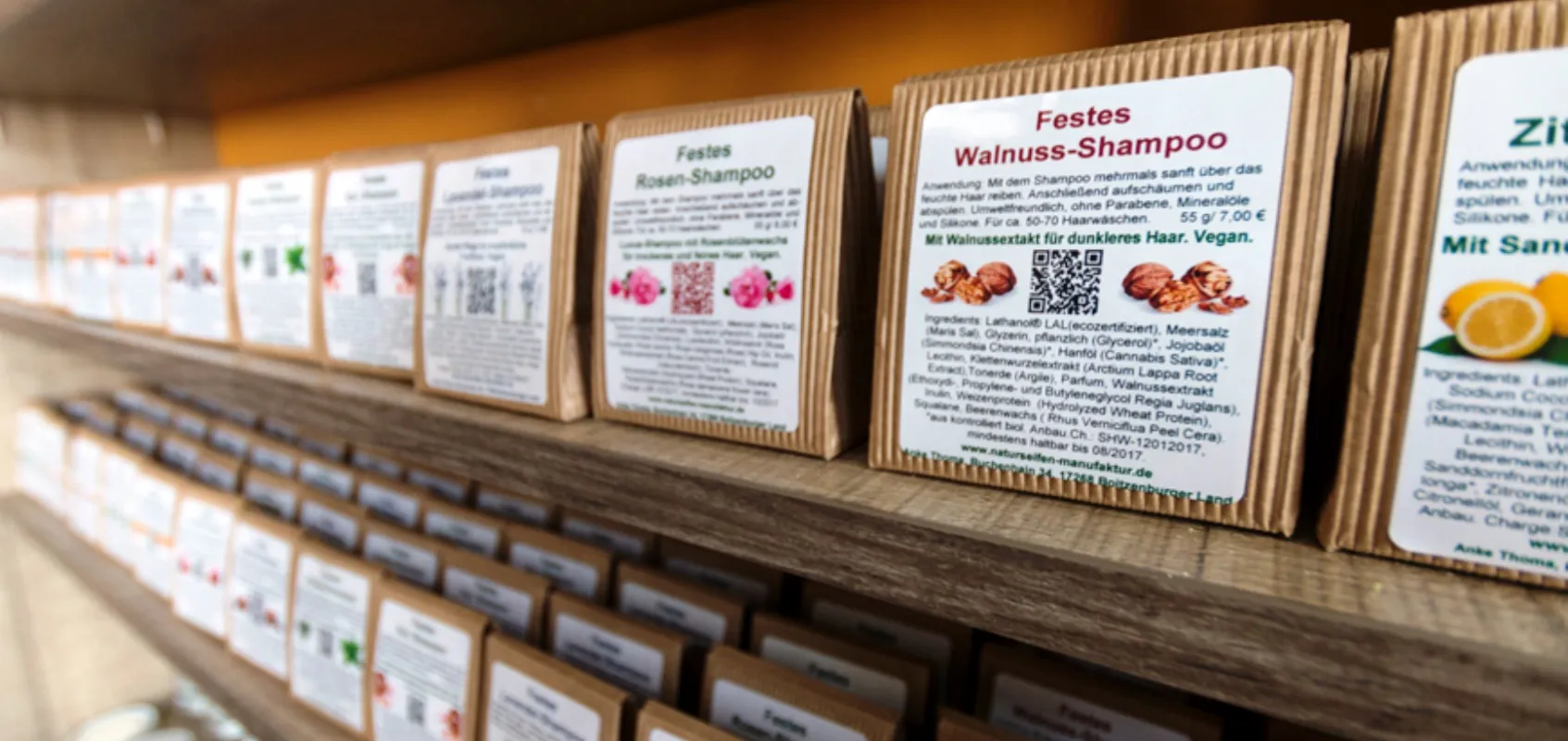 Regal mit handverpackten festen Shampoos in brauner Papierverpackung. Die Vorderseiten der Verpackungen zeigen die Inhaltsstoffe und Beschreibungen, darunter Walnuss- und Rosen-Shampoo.